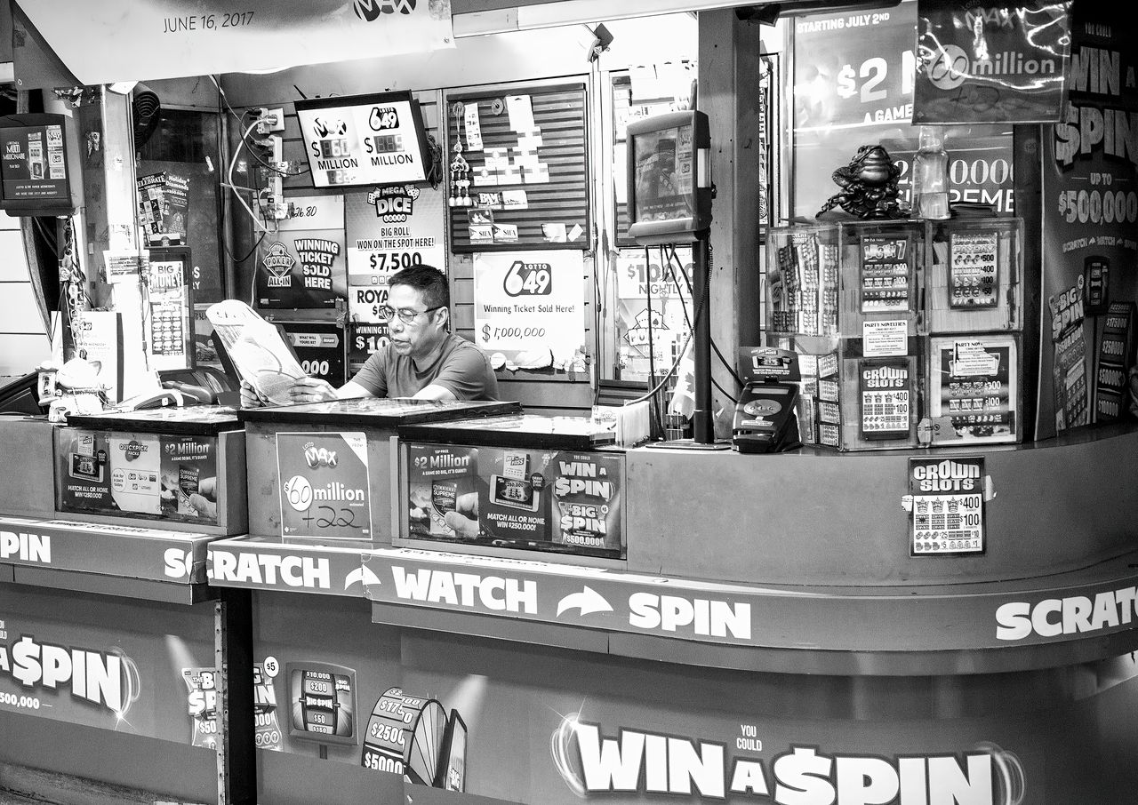 services-01 A man reads at a busy lottery ticket booth filled with signs and advertisements.